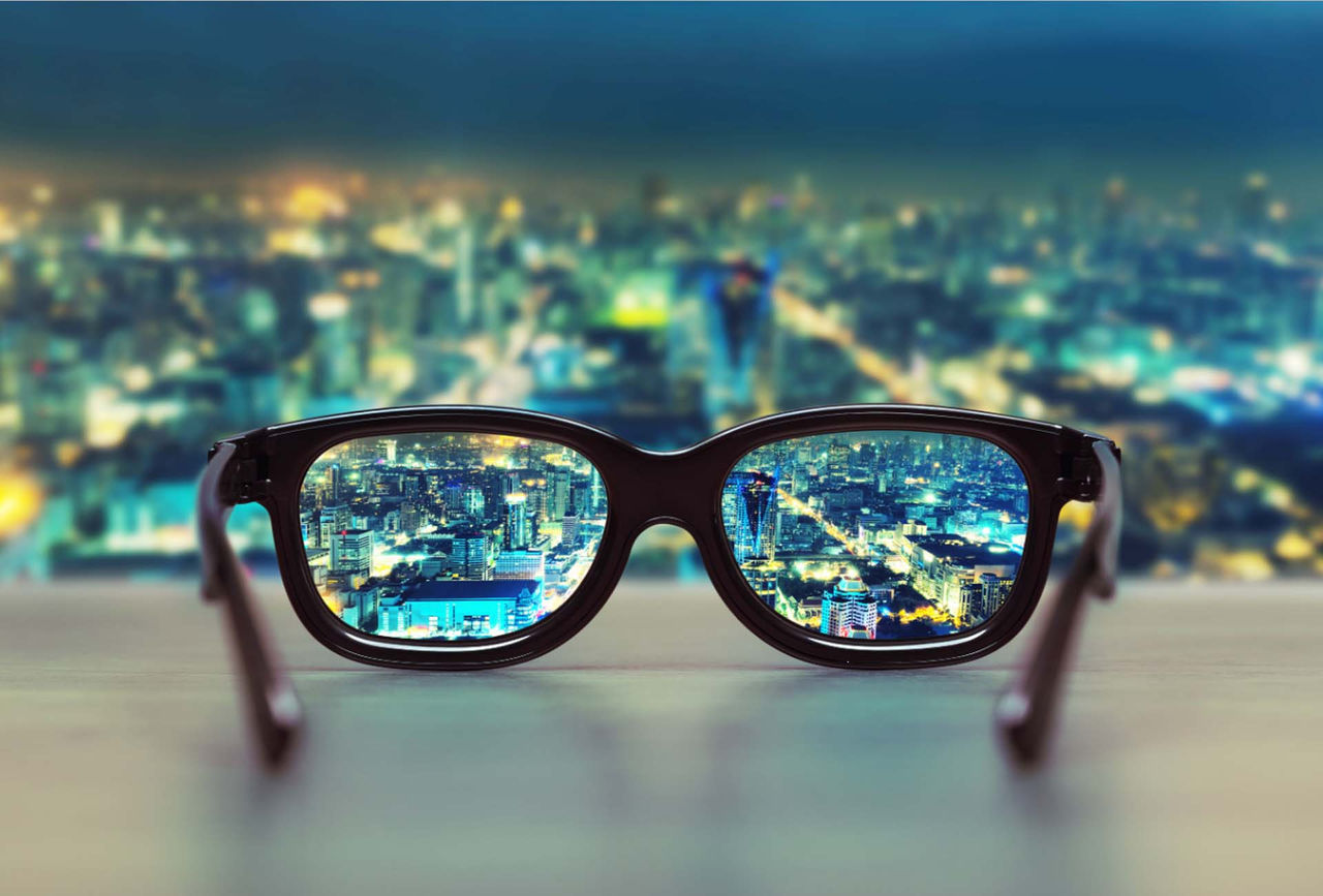 cityscape at night focused by a pair of glasses sitting on a polished wood surface